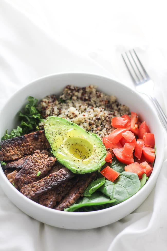 tempeh bacon, spinach, quinoa, kale tomatoes and half of avocado slice in a white bow with fork