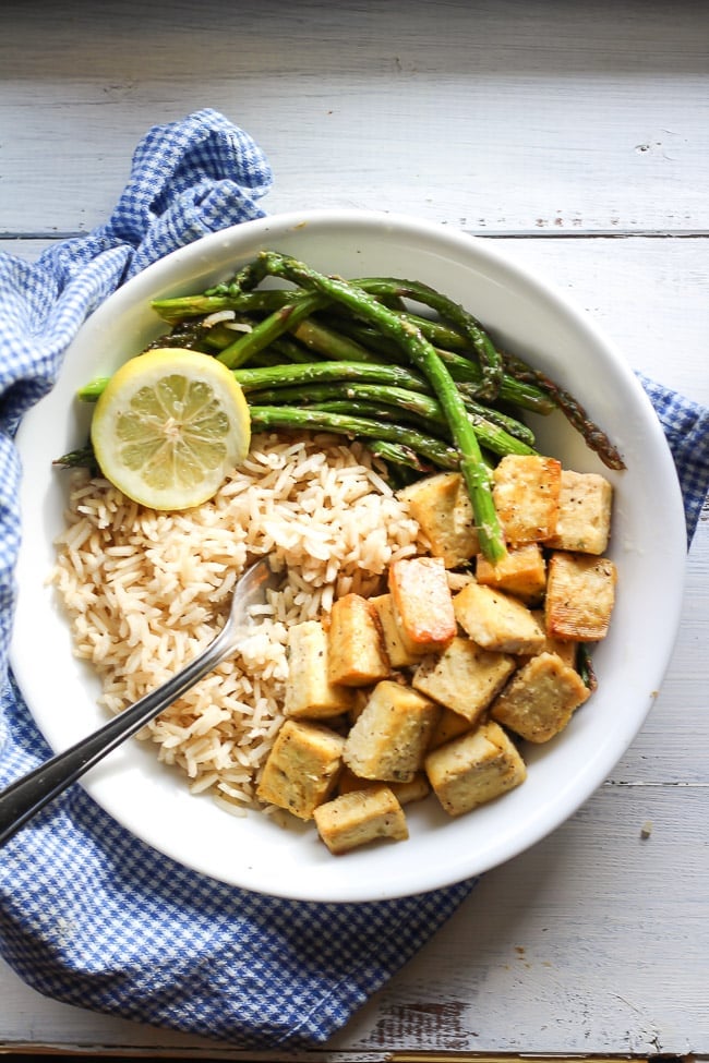 a picture of tofu, brown rice, and asparagus in a white bowl bowl with fork and lemon slice