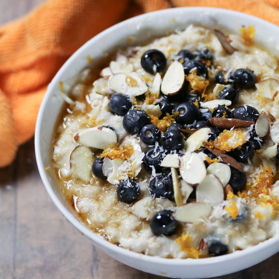 oats topped with blueberries, almonds, orange zest, and maple syrup in a white bowl
