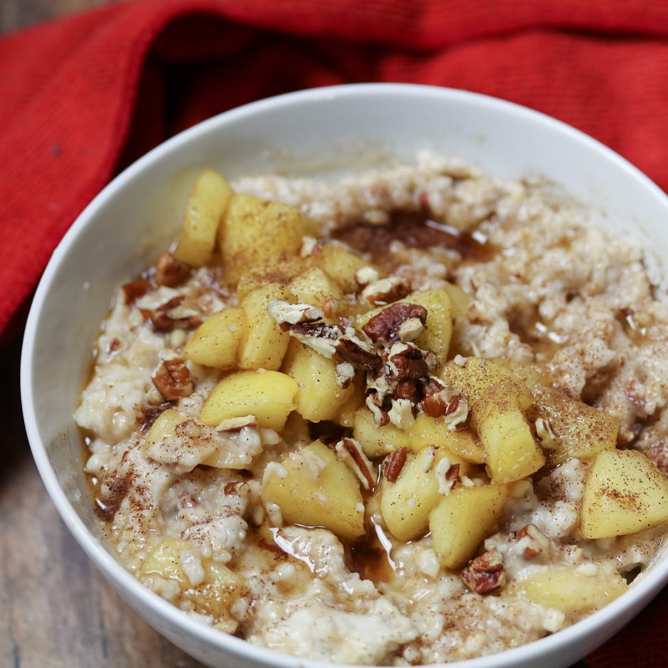 oats in a white bowl topped with cooked apples, pecans, and maple syrup