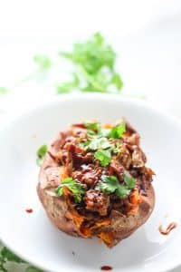 sweet potatoes on a white plate stuffed with lentils, mushrooms, and jackfruit topped with cilantro and bbq sauce