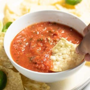 red vegan salsa in a white bowl with chip being dipped into it